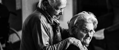Bild på en äldre kvinna som står bakom en annan äldre kvinna och håller sina händer på hennes axlar