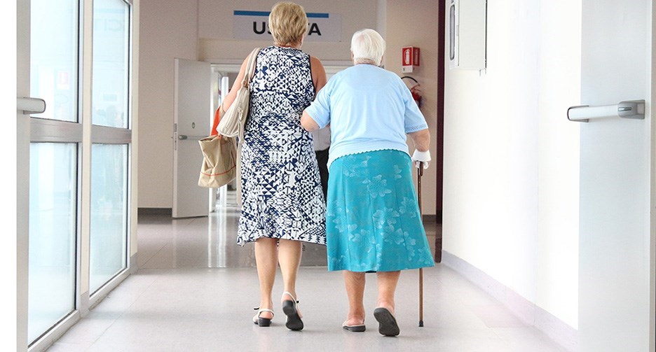 Två kvinnor sedda bakifrån som går i en sjukhuskorridor. En äldre kvinna stödjer sig på en käpp. En medelålders kvinna bär några väskor. Båda är sommarklädda i blå nyanser med klänning respektive kjol och t-shirt. Båda har sandaler.